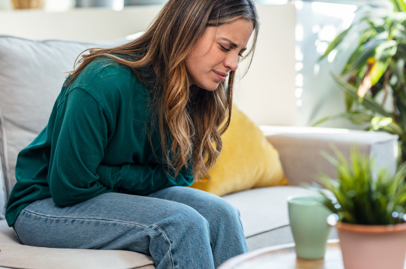 Young woman with cramps clutches her stomach and sits on the couch at home.
