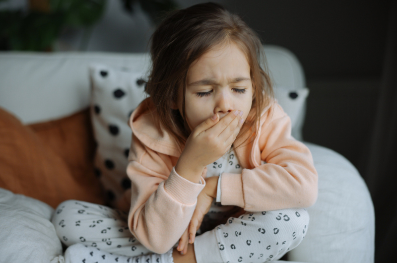 Little girl wearing pajamas sits on a sofa, coughing.