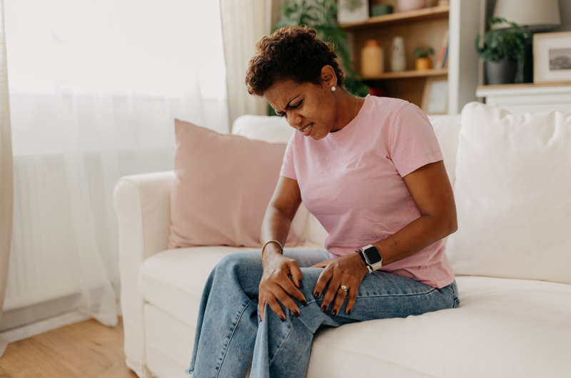 A woman in casual attire is seated on a white sofa, clutching her knee as she deals with leg pain.