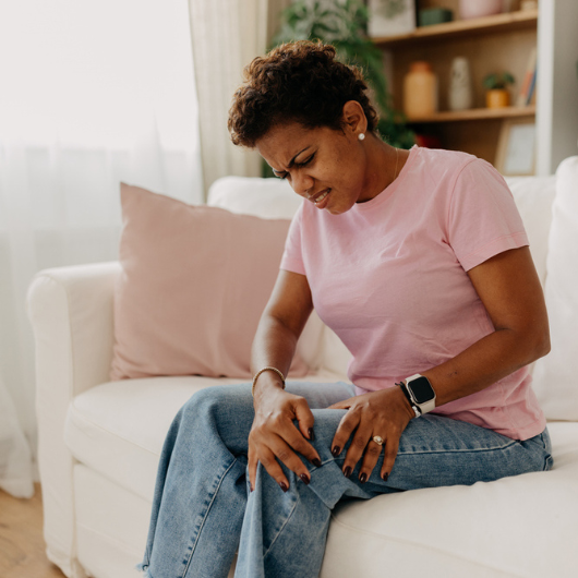 A woman in casual attire is seated on a white sofa, clutching her knee as she deals with leg pain.