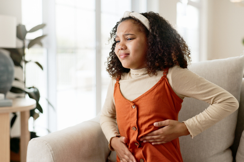 Young girl sits on a sofa at home clutching her stomach in pain.