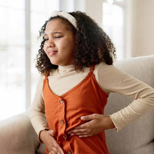 Young girl sits on a sofa at home clutching her stomach in pain.