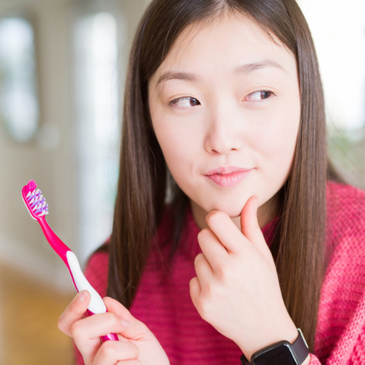 An Asian woman is deep in thought while holding a toothbrush.