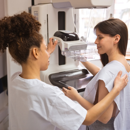 Female doctor talking to young woman during a mammogram in an examination room.