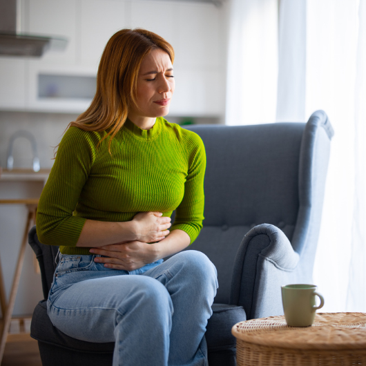 A young woman sits on a couch and clutches her abdomen in pain.  