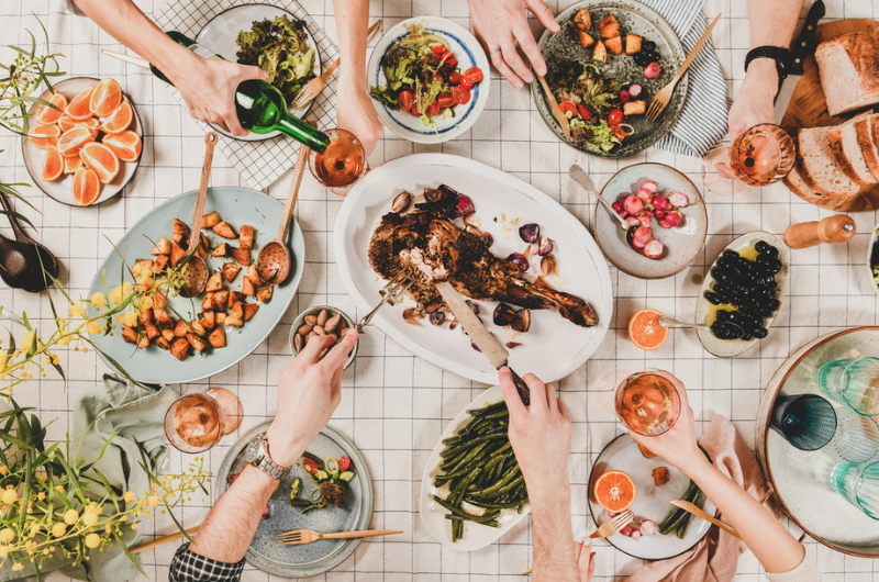Flat-lay of hands of people eating a Mediterranean diet spread of roasted lamb shoulder, salads, vegetables, etc.