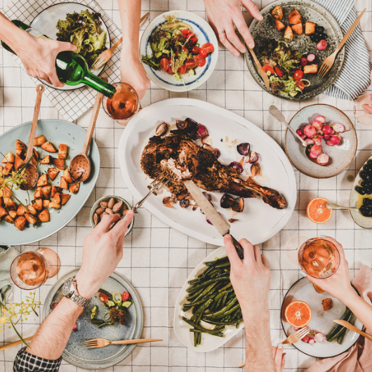 Flat-lay of hands of people eating a Mediterranean diet spread of roasted lamb shoulder, salads, vegetables, etc.