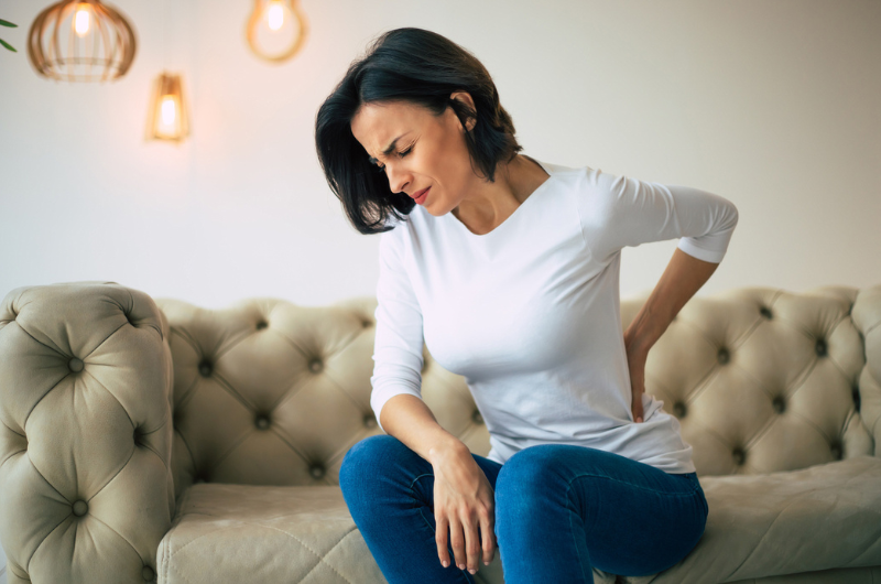 A woman in pain, sitting on a couch and holding her lower back with her left hand.