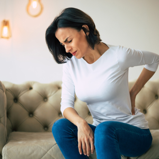 A woman in pain, sitting on a couch and holding her lower back with her left hand.