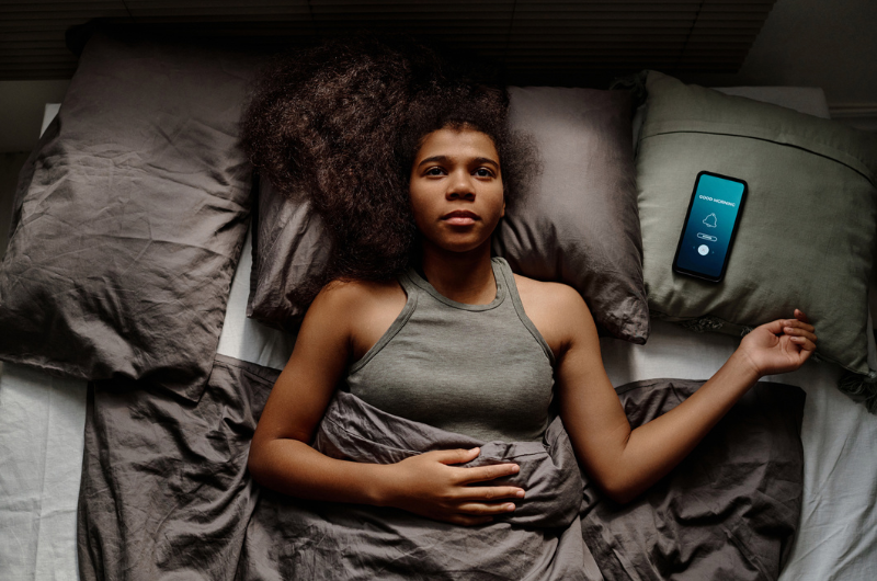 Top view of a worried African American female teenager with head on pillow while alarm clock on smartphone rings.