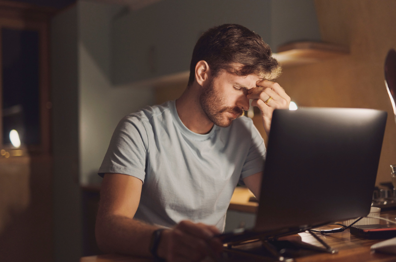  Man sitting in front of a desktop computer looking stressed, putting his hand on his forehead.   