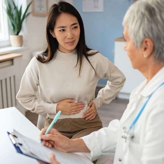 Female patient of Japanese ethnicity describes stomach pain to a Caucasian female doctor.