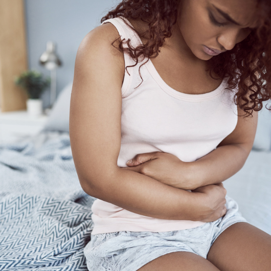 Young woman sits on the edge of her bed and clutches her lower stomach.