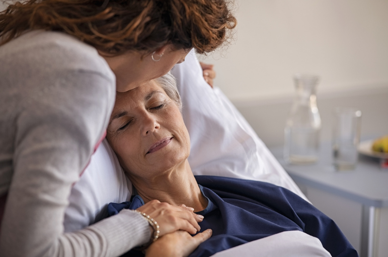 Close-up of adult daughter visiting and kissing her sick mother on the forehead while she rests in a hospital bed.