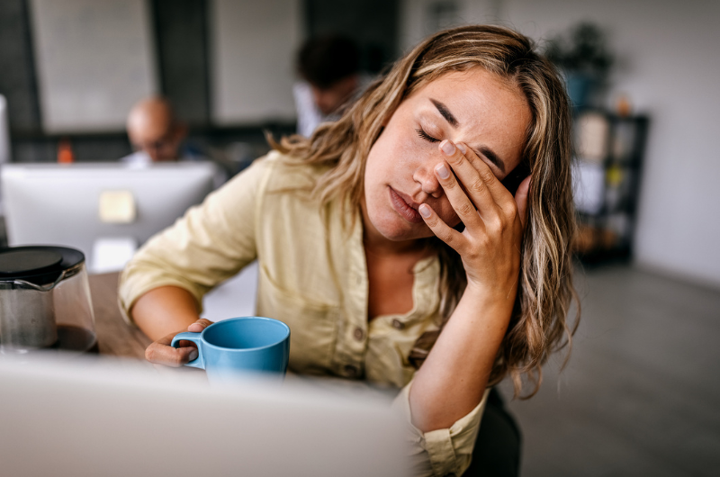 Exhausted young female entrepreneur drinking coffee and rubbing eyes.