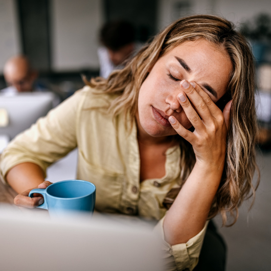 Exhausted young female entrepreneur drinking coffee and rubbing eyes.