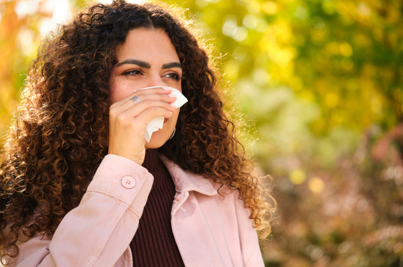 A young Caucasian woman with curly hair blows her nose into a tissue during autumn.