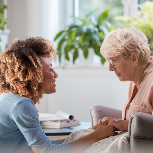 African American female home nurse talks with a senior woman, sitting in the living room and holding her hands.