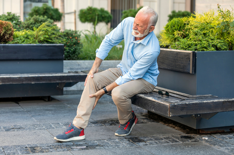 Man with white hair sits outside on a bench and holds his leg, which is causing him pain.