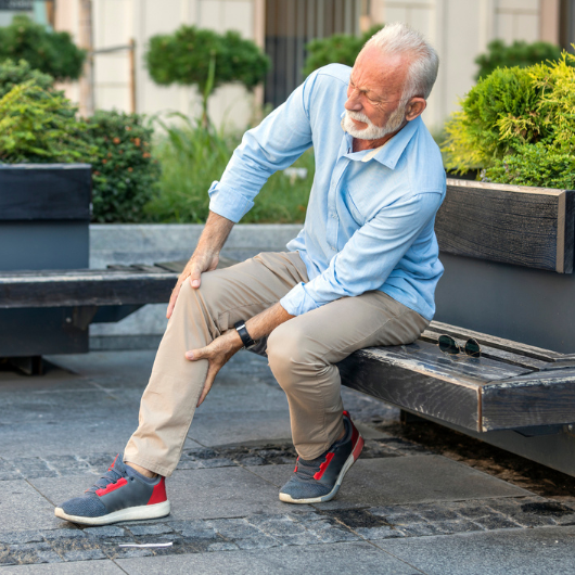 Man with white hair sits outside on a bench and holds his leg, which is causing him pain.