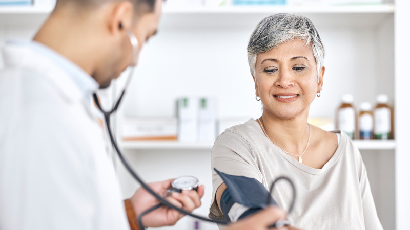 Image shows a doctor checking a woman's blood pressure in a clinical setting.