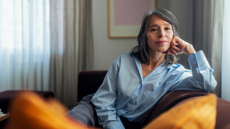 A calm, pensive woman sits comfortably on a sofa in a softly lit living room.