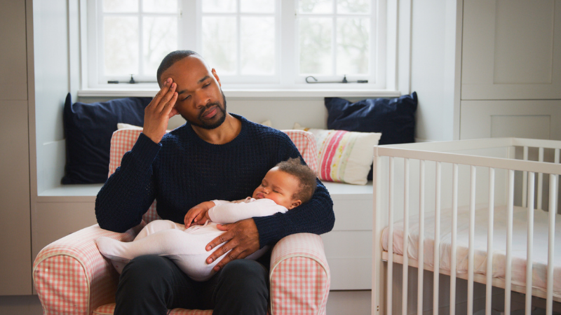 Stressed new father sitting in a chair holding a sleeping baby girl.