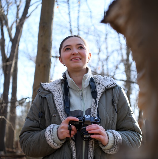 Young woman, Caitlyn, stands in the woods holding a camera, looking out and smiling.