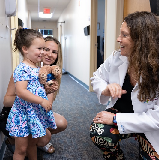 Toddler Maggie Hennessey smiling in the doctor's office hallway, with her mom and doctor.