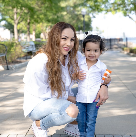 Marisol and her young daughter standing in the park smiling together.