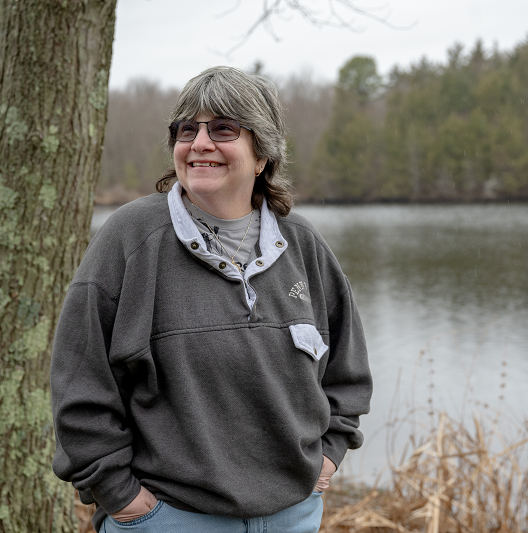 Woman, Elly Levine, standing in front of a lake smiling.