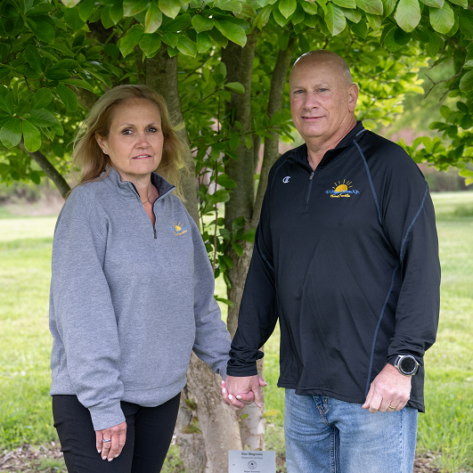 Stu and Michele Schulman standing outside, holding hands, under a tree.