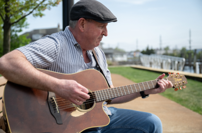 A man strums a guitar while sitting next to a river.
