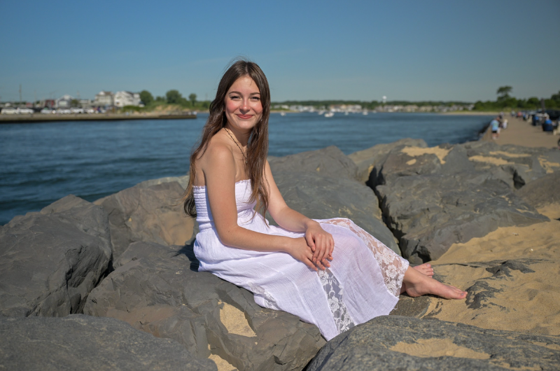 A teen girl sits on a large rock at the edge of a body of water.