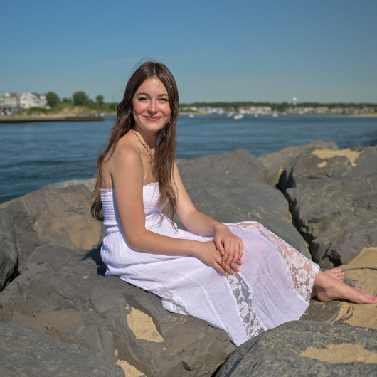 A teen girl sits on a large rock at the edge of a body of water.