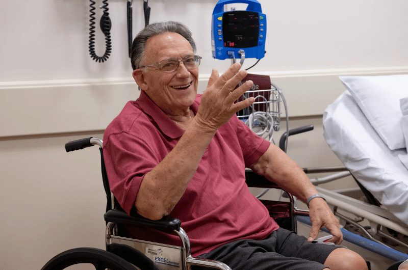 79-year-old Anthony Scuorzo smiles at the camera from a wheelchair in a patient room.