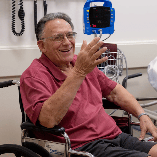 79-year-old Anthony Scuorzo smiles at the camera from a wheelchair in a patient room.