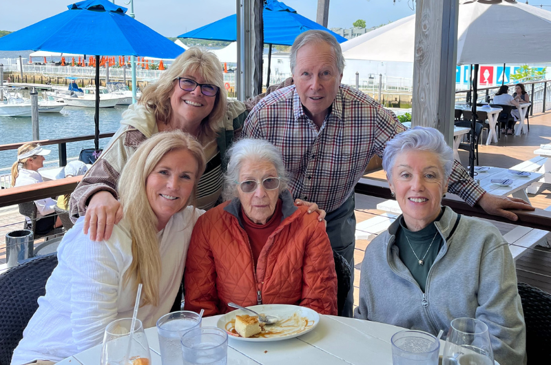 While dining outdoors, an elderly woman and her family smile at the camera.