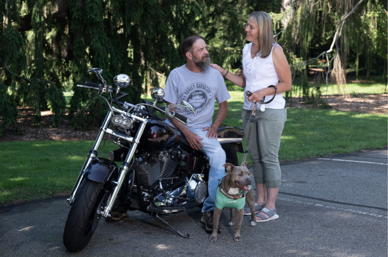 William Kopsco sits on his motorcycle while Jules Kopsco stands near and places her hand on his shoulder.