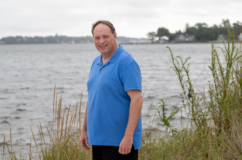 Standing in front of a scenic body of water, Joe Keenan smiles at the camera.