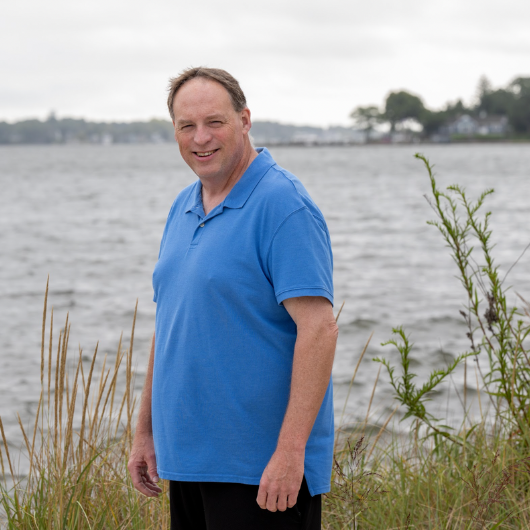 Standing in front of a scenic body of water, Joe Keenan smiles at the camera.