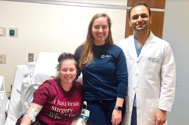 Selena Campione, 36, sits in a patient bed with brain surgeon, Nitesh Patel, M.D., standing beside her.