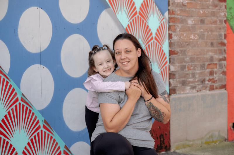 With her young daughter standing behind her, Lisa Ruding kneels and smiles.