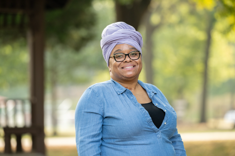 Woman smiles at the camera with greenery behind her.
