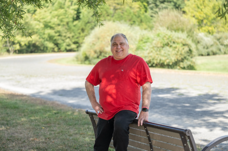 Nick Pattakos leans against a bench in a park while smiling at the camera.
