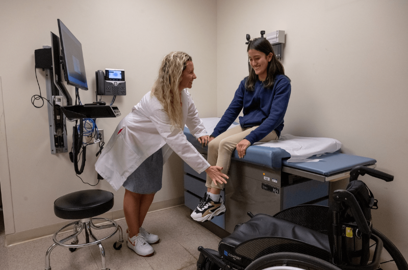 Teenager Melanie Mejia sits on an exam table as her doctor, a blonde female, talks to her and smiles.