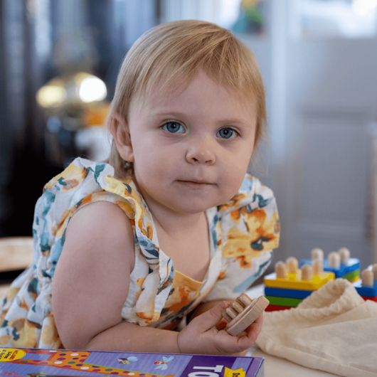 Toddler Marigold Mackey leans over a table covered in toys while looking at the camera.