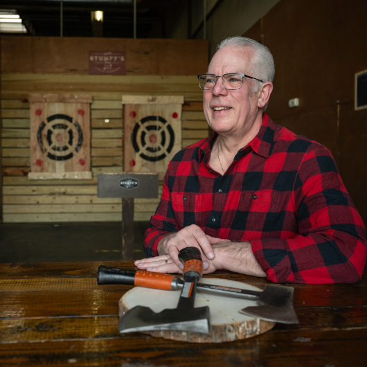With a background of an ax-throwing lane, Scott Knox looks off camera and smiles.