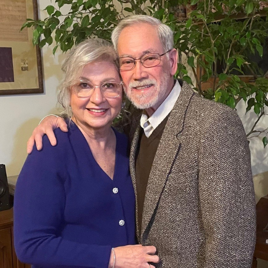 Breast cancer survivor Nora Ambros smiles at the camera while sitting next to her husband.