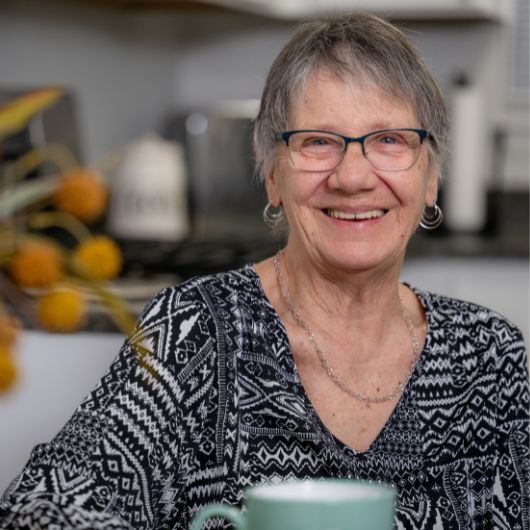 74-year-old Cecelia Lucas sits with a cup of coffee and smiles at the camera.